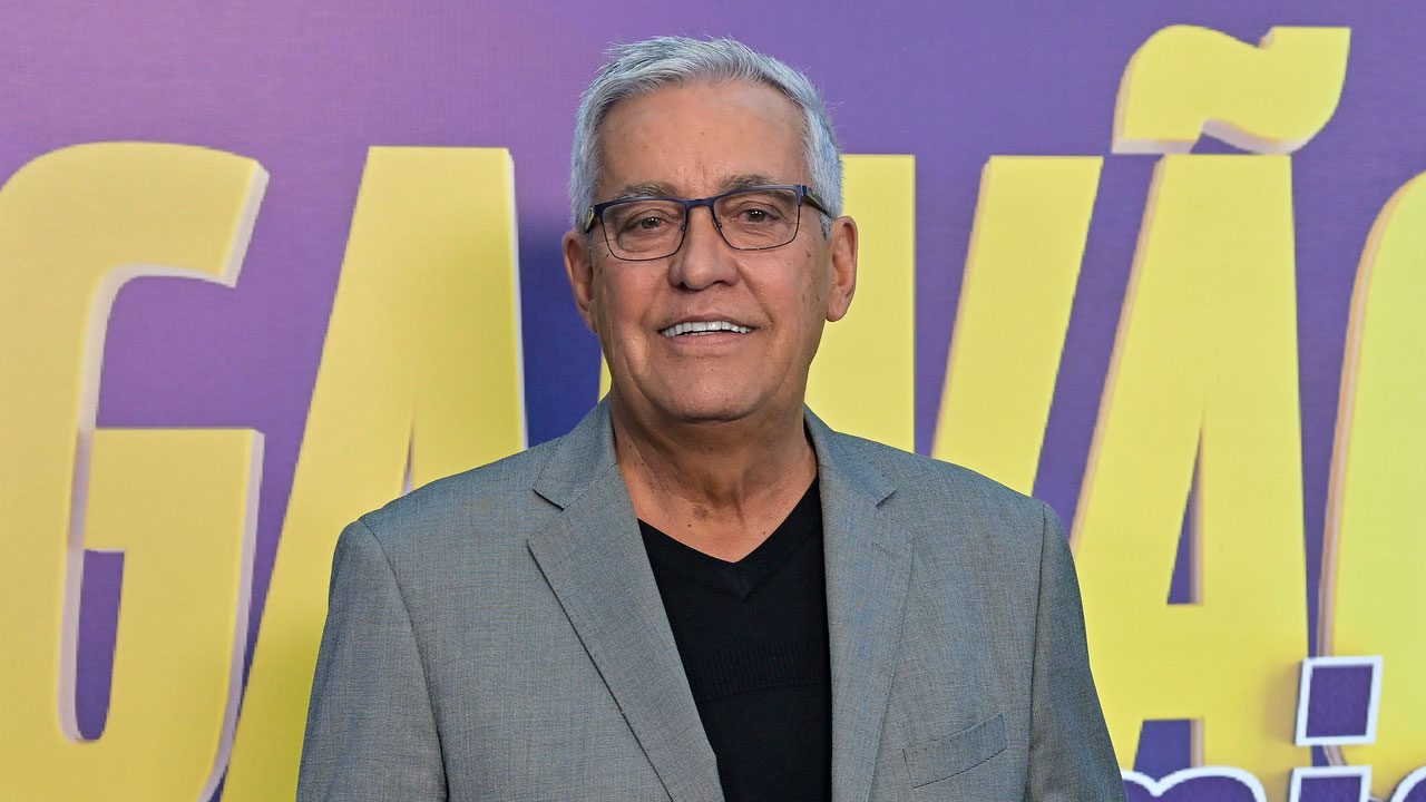 Jornalista Mauro Naves sorrindo, de óculos e blazer cinza, em evento de lançamento do Galvão e Amigos