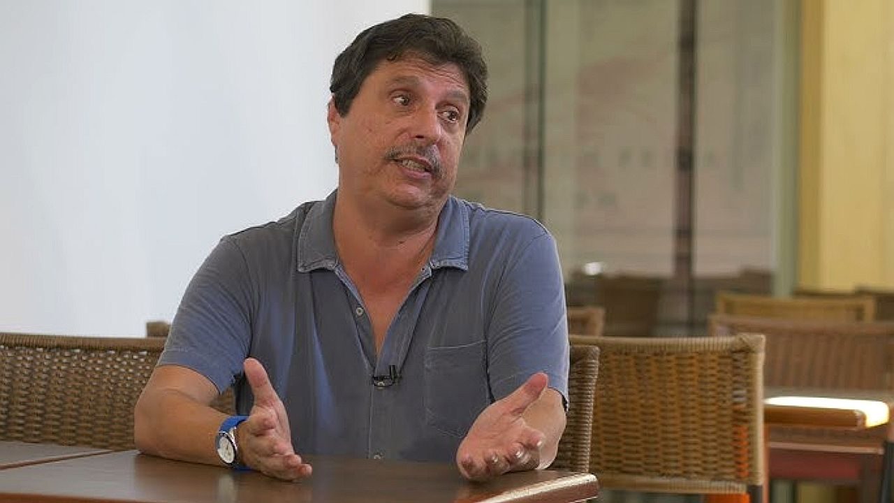 Mauro Wilson durante entrevista com microfone de lapela sentado à mesa e gesticulando enquanto fala sobre televisão brasileira