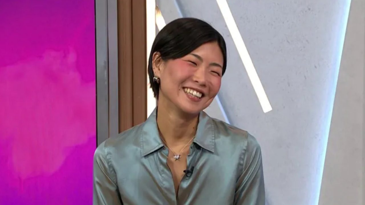 May Hong sorri durante entrevista em estúdio colorido vestindo camisa acetinada azul clara e colar prateado com pingente em formato de estrela