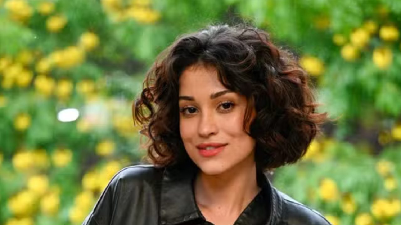 Mulher com cabelo curto e cacheado, vestindo jaqueta escura, olha para a câmera com leve sorriso. Ao fundo, há vegetação verde com flores amarelas desfocadas, sugerindo um ambiente externo e natural.