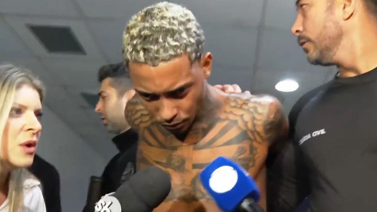 MC Poze é escoltado por policiais civis e cercado por repórteres em corredor de delegacia