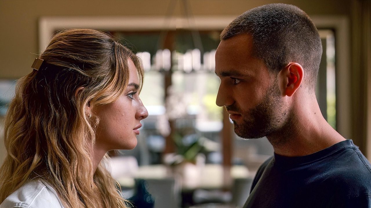 Um homem e uma mulher estão frente a frente, muito próximos, trocando um olhar intenso. Ela tem cabelos longos com mechas claras presos parcialmente atrás e veste uma blusa clara. Ele tem cabelo bem curto e barba aparada, usando uma camiseta escura. O ambiente parece ser interno, com luz natural entrando por janelas ao fundo. A expressão de ambos sugere uma conversa séria ou um momento emocionalmente carregado.