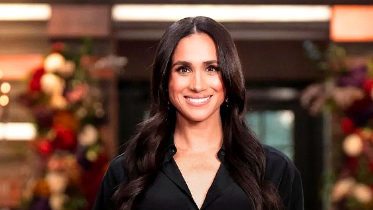 Meghan Markle sorri usando camisa preta em imagem promocional gravada em ambiente interno decorado com flores