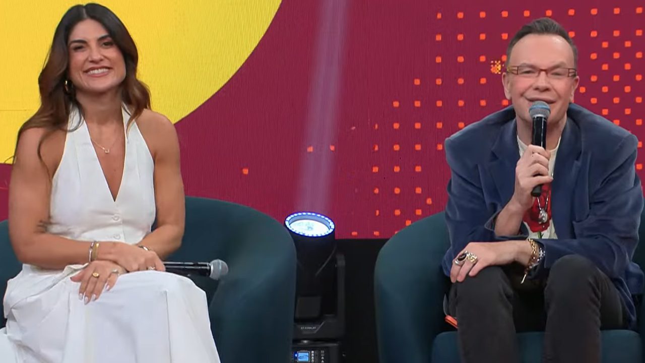 A imagem mostra Pâmela Lucciola e Felipeh Campos sentados em poltronas no palco do Melhor da Noite. A mulher, vestindo um macacão branco, sorri para a câmera enquanto segura um microfone no colo. O homem ao lado dela, usando óculos e um blazer azul, também sorri e segura um microfone, parecendo estar falando. O fundo colorido e as luzes de estúdio indicam um ambiente descontraído, típico de um programa de entretenimento