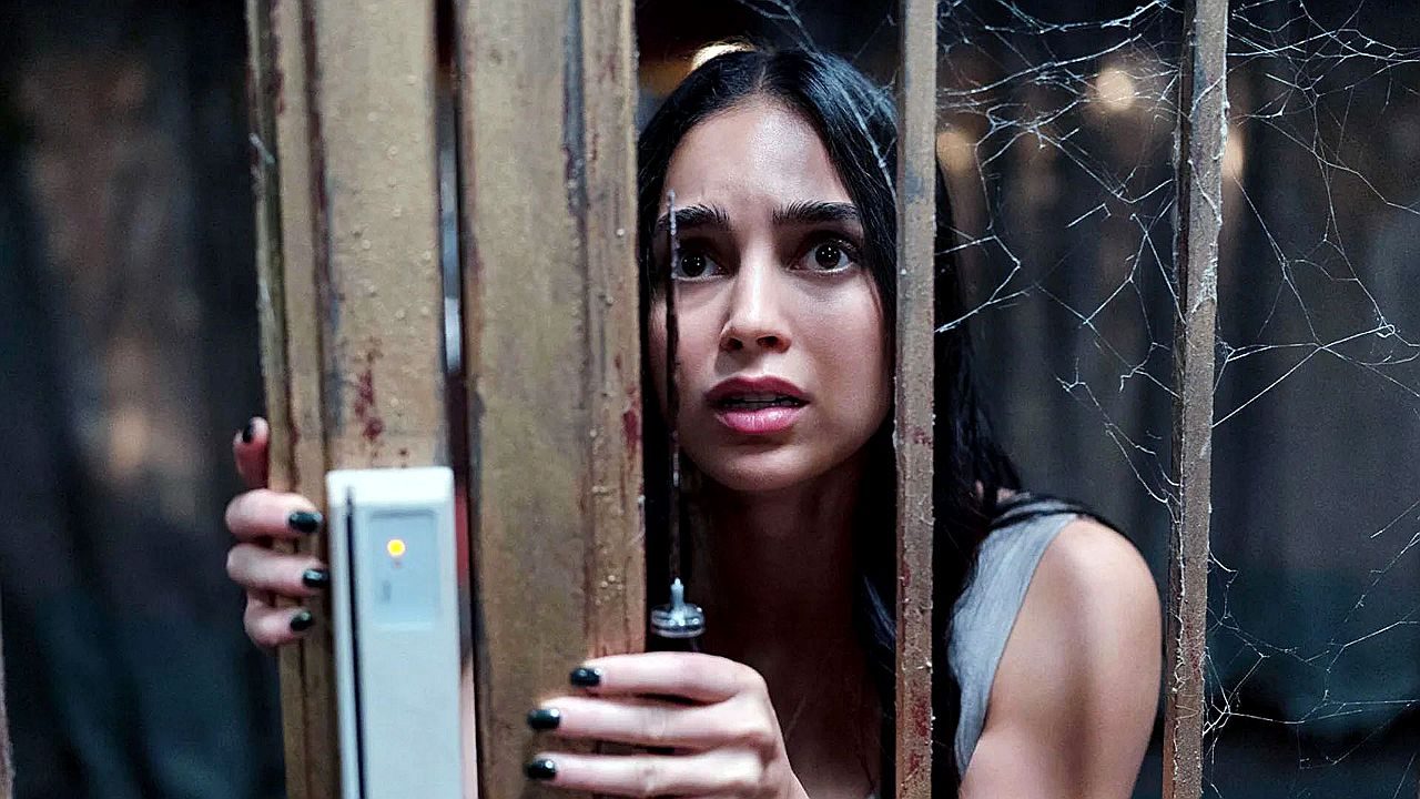 Mulher assustada segura porta de elevador antigo cercada por teias de aranha em cena de suspense