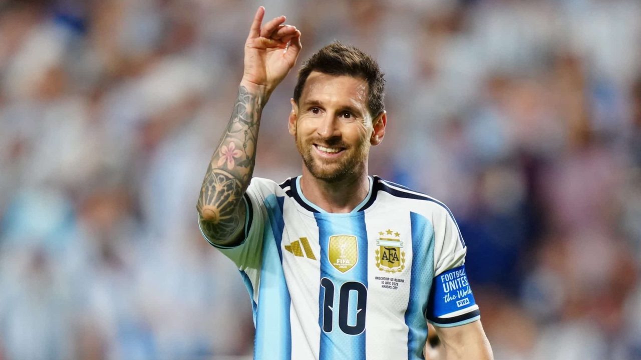 Lionel Messi, com a camisa 10 da Argentina, sorri e ergue a mão durante partida, com braçadeira de capitão e torcida desfocada ao fundo