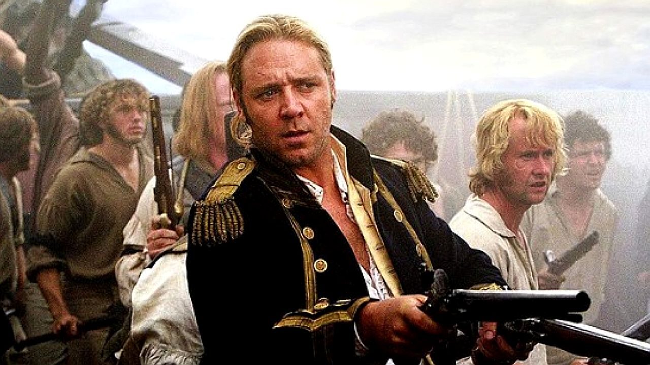 Homem usando uniforme militar antigo, segurando uma arma, em meio a uma cena de batalha com vários soldados ao redor e fumaça no ambiente.