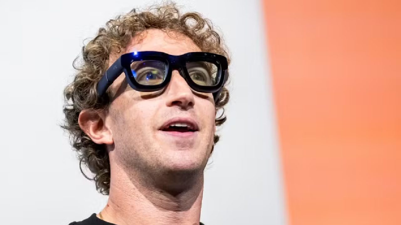 Homem jovem de pele clara e cabelos cacheados loiros usa óculos grandes de armação preta brilhante; ele aparece em close, olhando ligeiramente para cima, diante de um fundo dividido entre branco e laranja