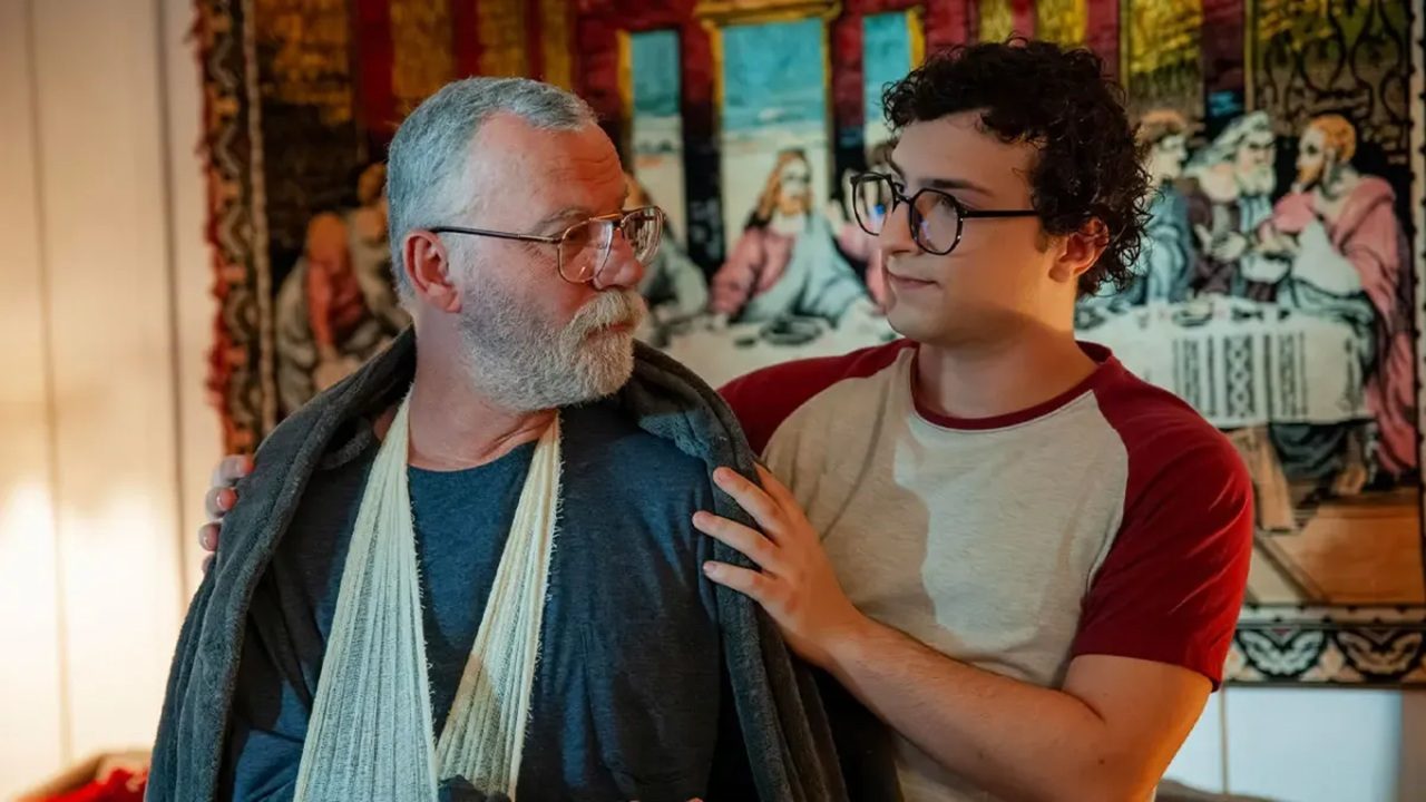 Homem mais velho com o braço enfaixado recebe apoio de um jovem que o segura pelos ombros; ambos se encaram com expressão afetuosa, em ambiente interno decorado com tapeçaria religiosa ao fundo.