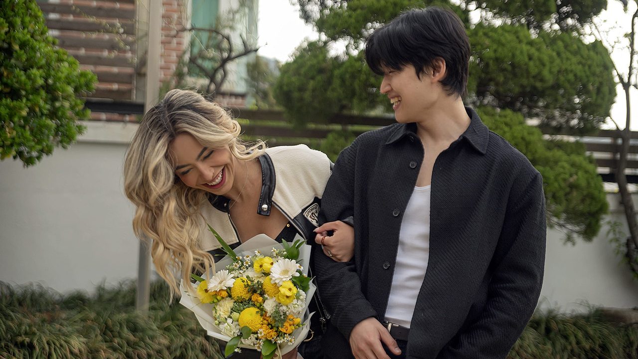 Duas pessoas caminham sorrindo ao ar livre enquanto se olham, uma delas segura um buquê de flores amarelas e brancas e a outra segura seu braço, com árvores e muro ao fundo