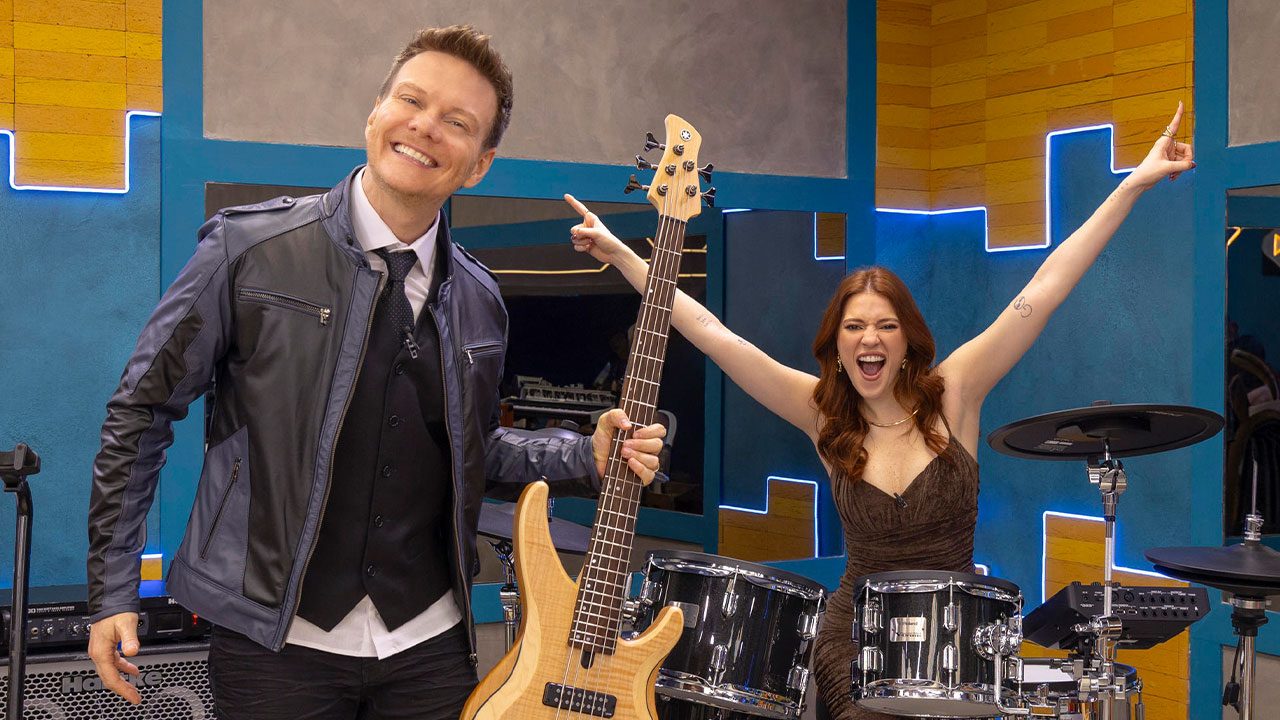 Michel Teló sorrindo e segurando baixo ao lado de Ana Clara comemorando atrás de bateria na casa do Estrela da Casa