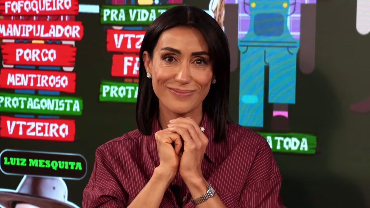 Michelle Barros sorrindo, com as mãos cruzadas, no estúdio do programa e tela de LED com termos como "Fofoqueiro" e "Porco"