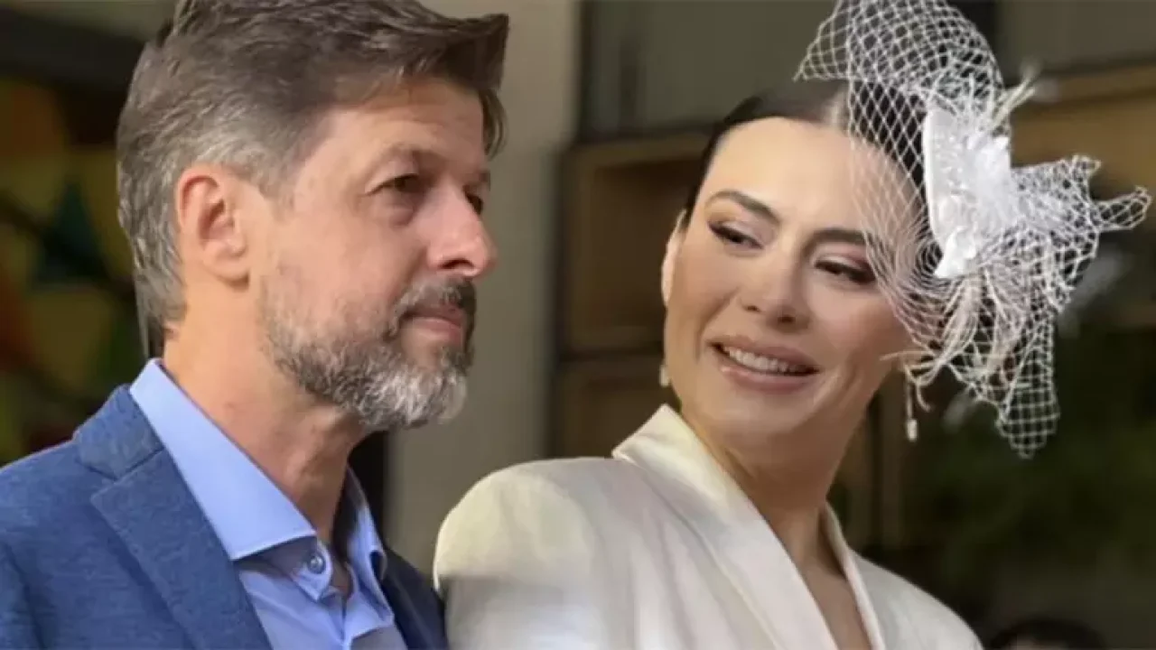 Michelle Loreto sorri para Alexandre Mattoso durante cerimônia, usando uma roupa branca e traje elegante