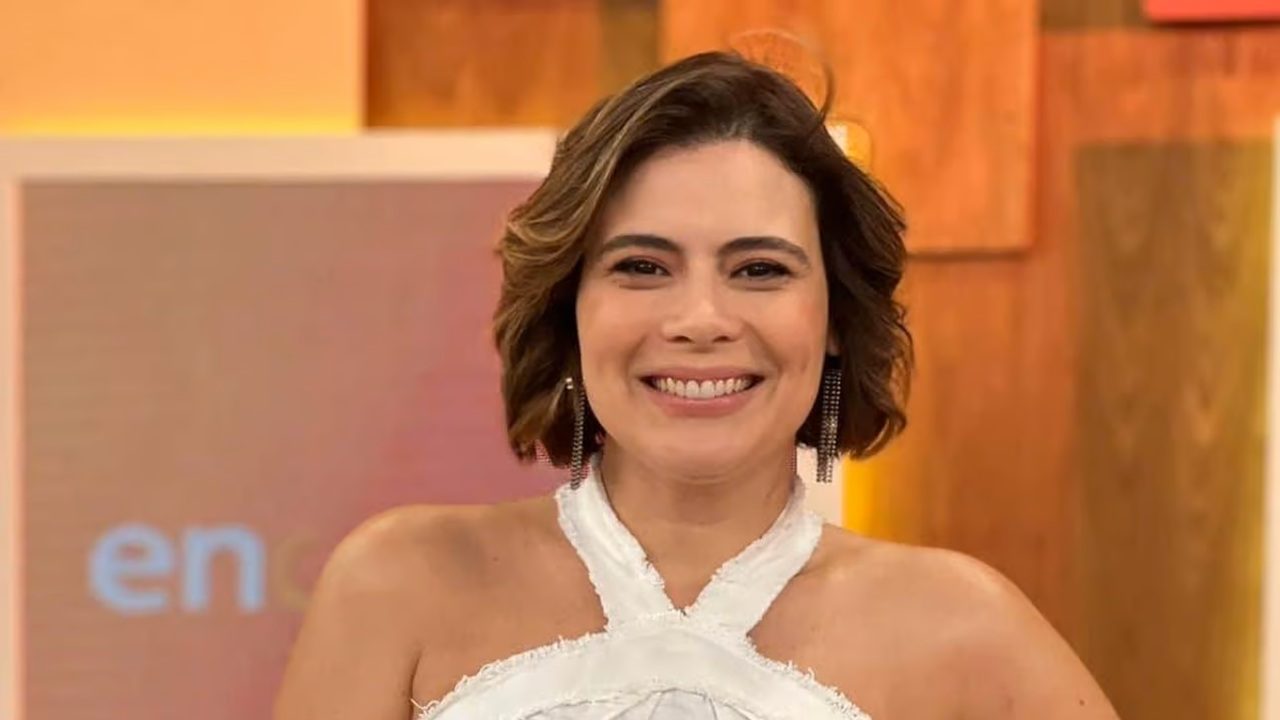 A imagem mostra uma mulher sorridente, com cabelo curto e castanho, vestindo um top branco com gola alta cruzada. Ela está em um estúdio de televisão, com um fundo colorido em tons de laranja e marrom, que exibe parte de um logotipo desfocado