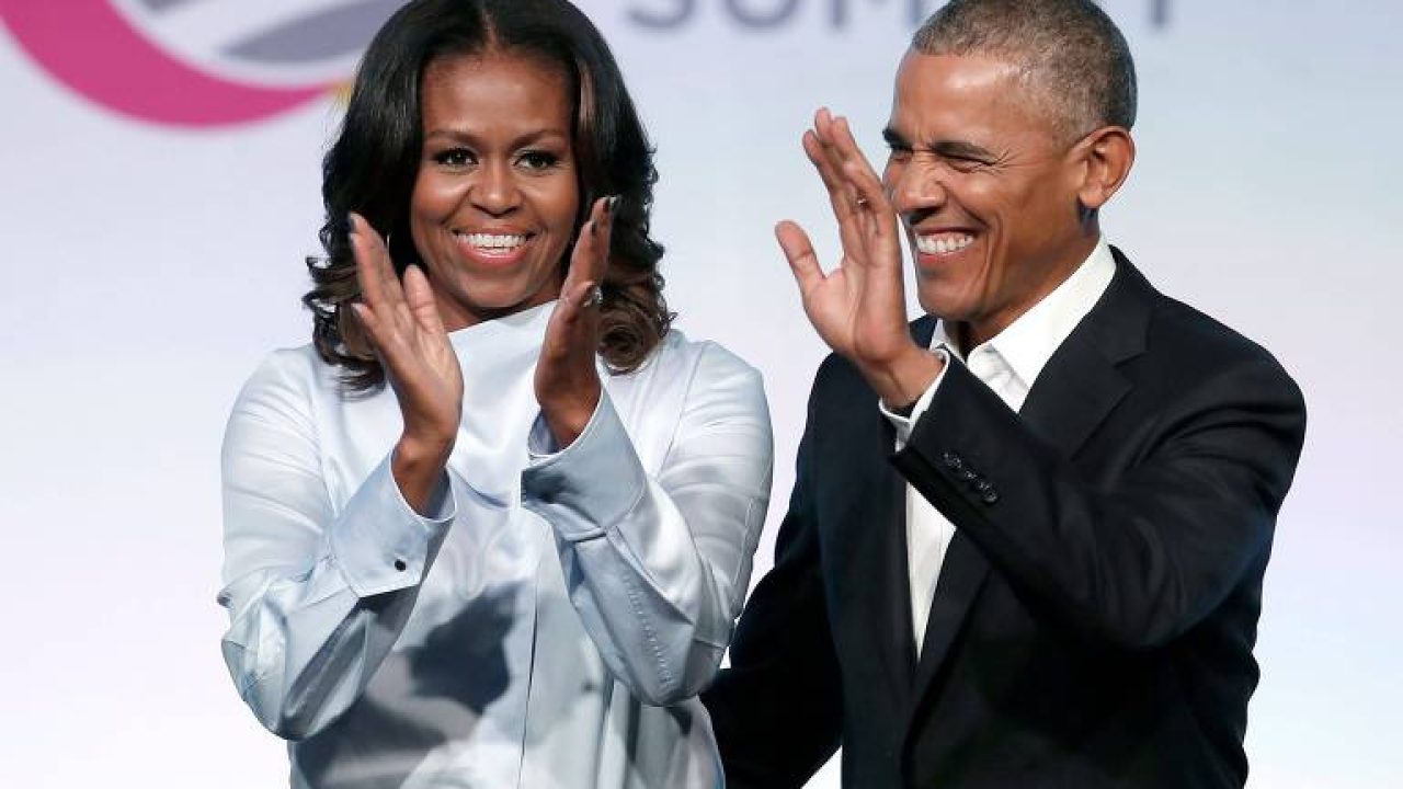 Michelle Obama e Barack Obama aplaudindo e sorrindo durante evento com palco ao fundo