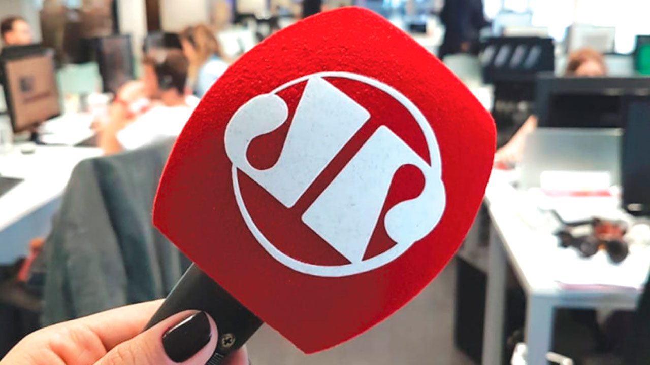 Microfone com logotipo da Jovem Pan sendo segurado em redação com pessoas ao fundo