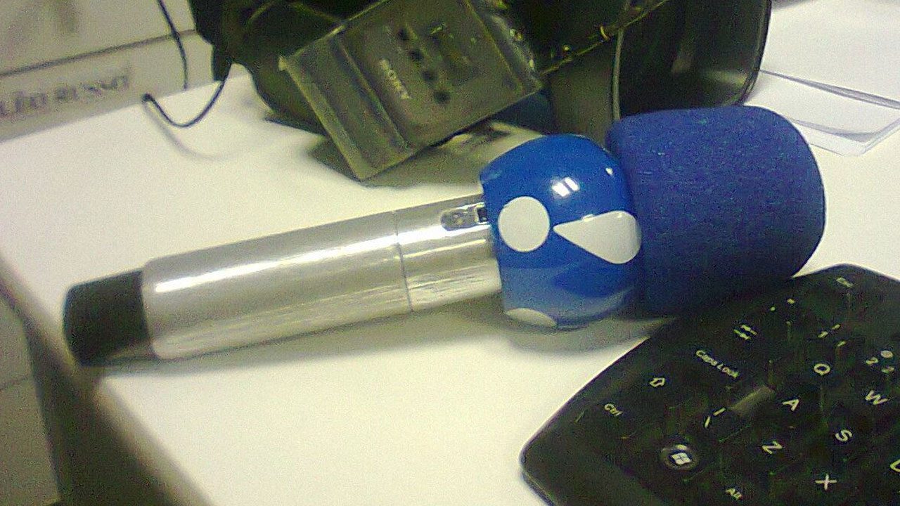 Microfone da RedeTV! com espuma azul e logo branco apoiado sobre mesa ao lado de teclado e câmera