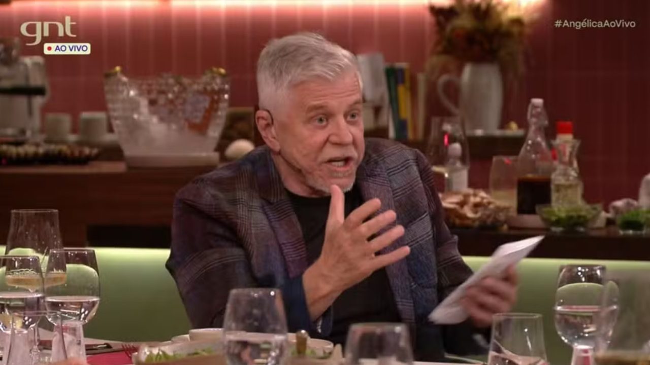 Miguel Falabella durante uma conversa em programa de TV, gesticulando com uma das mãos enquanto segura um papel, em mesa posta com taças e pratos, em ambiente elegante do canal GNT