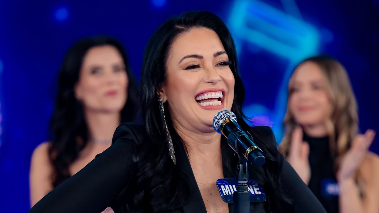 Milene Pavorô sorrindo ao microfone com crachá azul no Programa Silvio Santos com bailarinas do balé ao fundo