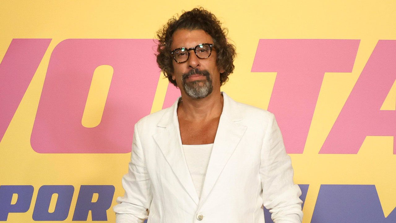 Milhem Cortaz com blazer branco em frente ao painel da novela Volta por Cima durante o lançamento da trama
