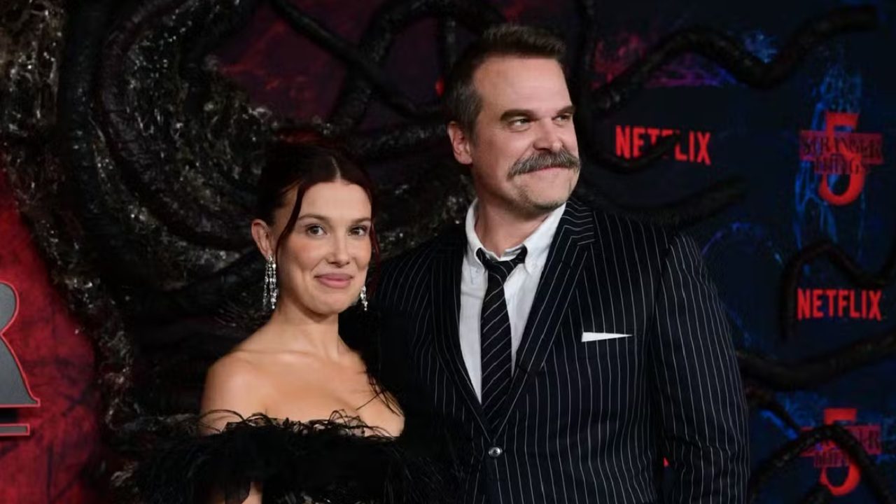 Dois atores posam sorrindo em evento da série Stranger Things, com o logotipo da produção e o nome da Netflix iluminados em vermelho ao fundo