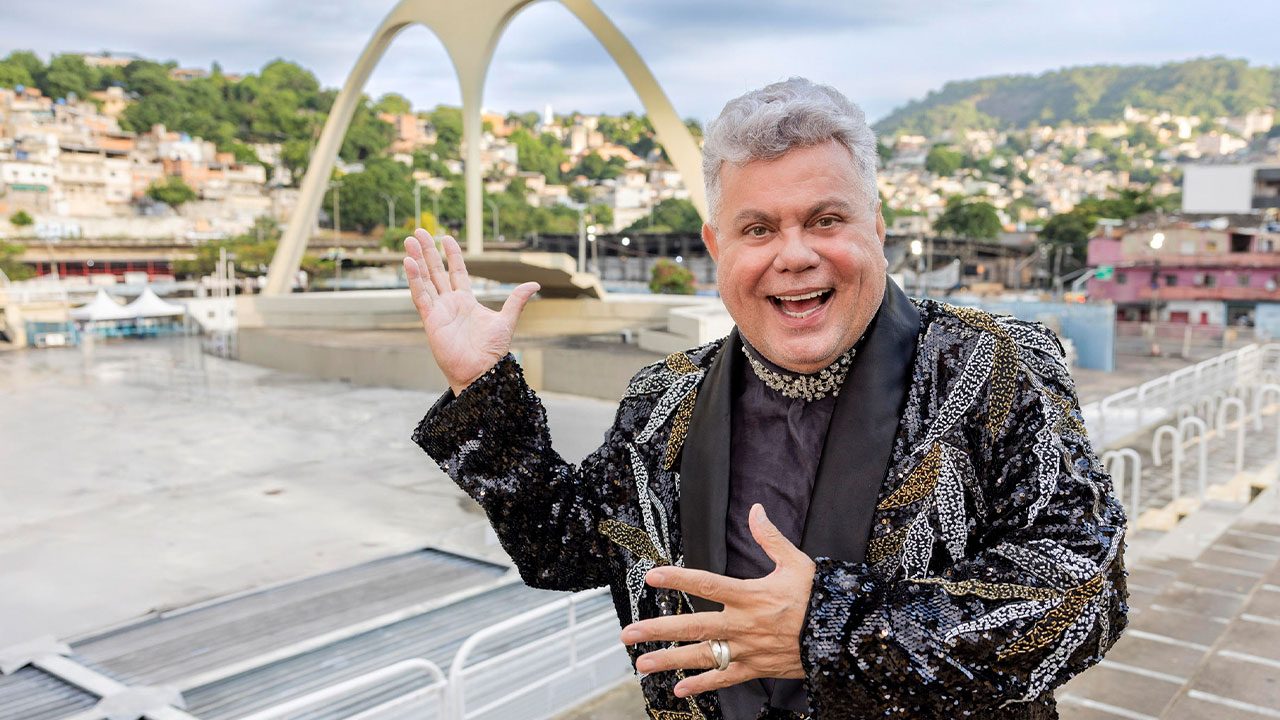 Milton Cunha sorri e gesticula no Sambódromo da Marquês de Sapucaí, o carnavalesco usa paletó preto com paetês e detalhes dourados, ao fundo a Apoteose