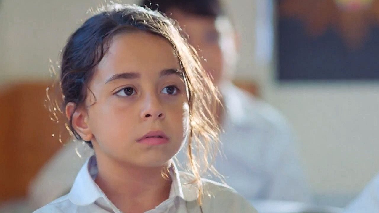 Menina de uniforme branco observa algo com expressão séria e atenta em sala iluminada com pessoas desfocadas ao fundo