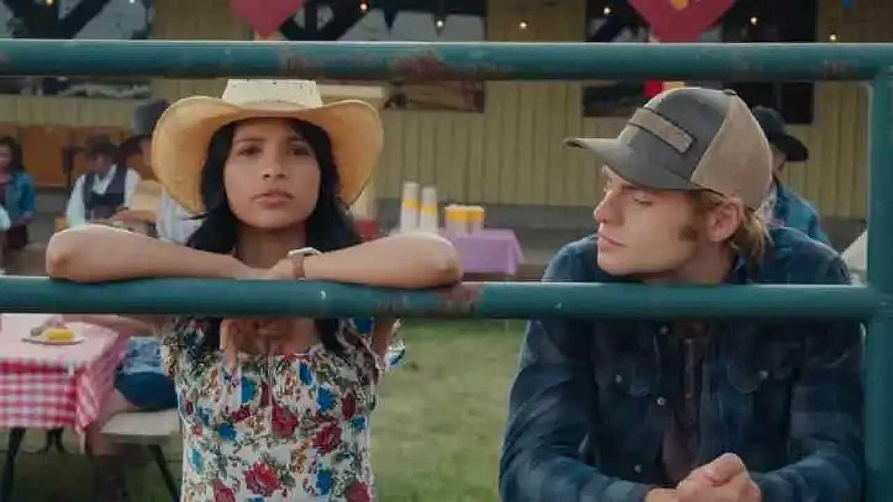Casal conversa apoiado em cerca durante festa rural em cena de série adolescente
