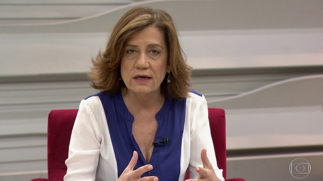 A jornalista Míriam Leitão aparece em estúdio da TV Globo durante participação em um programa da GloboNews. Sentada em uma poltrona vermelha, ela usa uma blusa branca com detalhes em azul e gesticula com as mãos enquanto analisa o cenário político e econômico. Com expressão séria, Míriam mantém seu tom característico de comentarista experiente.