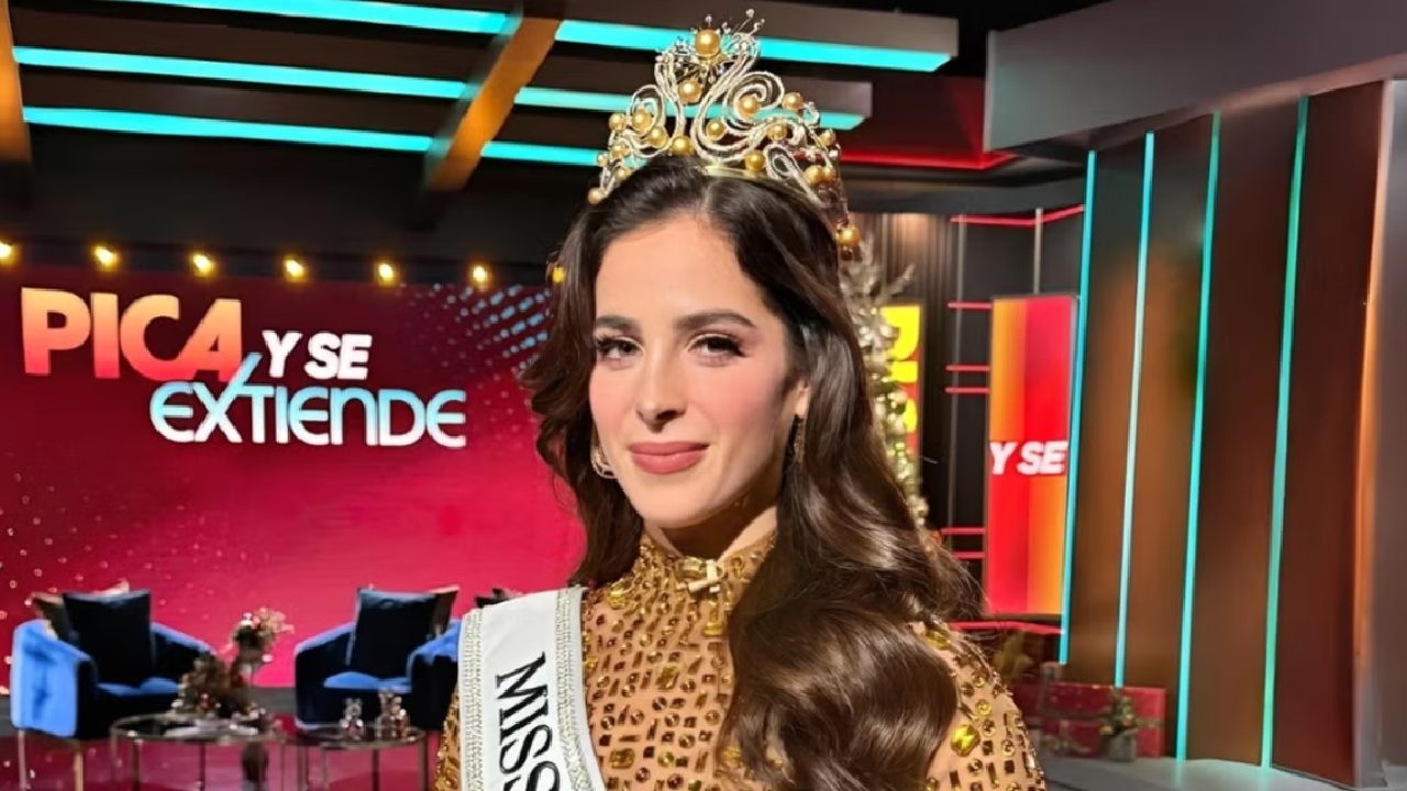 Mulher usando uma coroa ornamentada e uma faixa de concurso de beleza, posando em um estúdio de televisão colorido, com luzes e cenografia ao fundo.