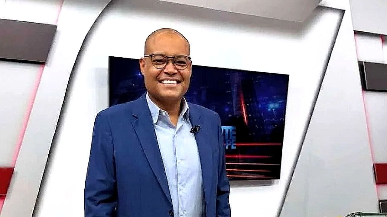 Homem de terno sorri em estúdio de telejornal com tela ao fundo exibindo cenário urbano