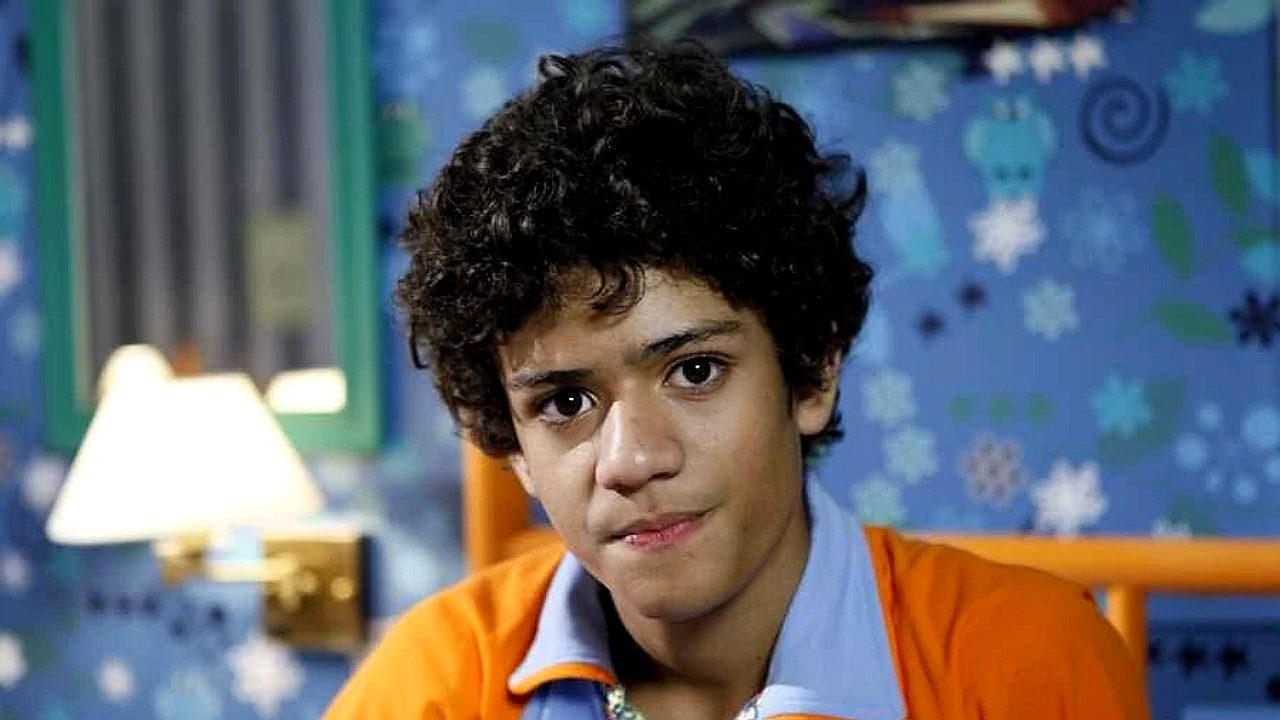 Menino de cabelo cacheado encara a câmera com expressão séria em quarto com parede azul e desenhos ao fundo