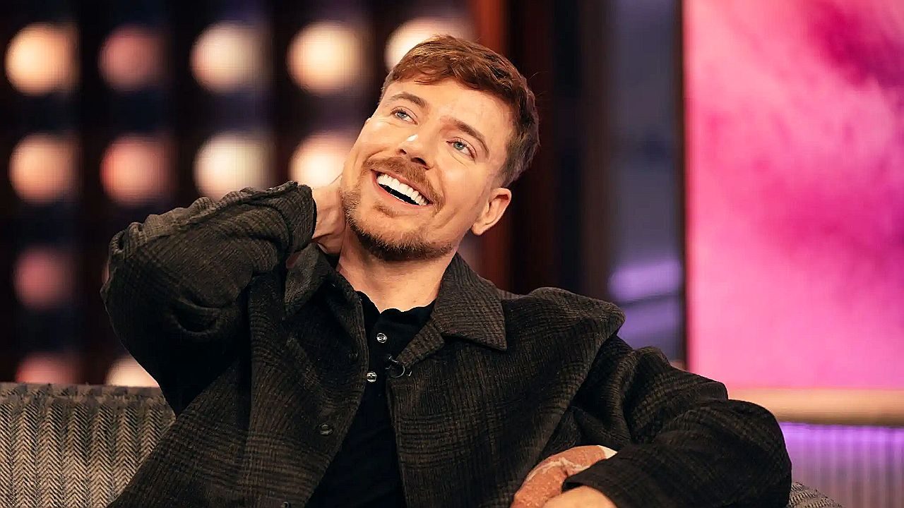 Ator sorri e leva a mão ao pescoço durante entrevista em programa de TV com cenário iluminado ao fundo