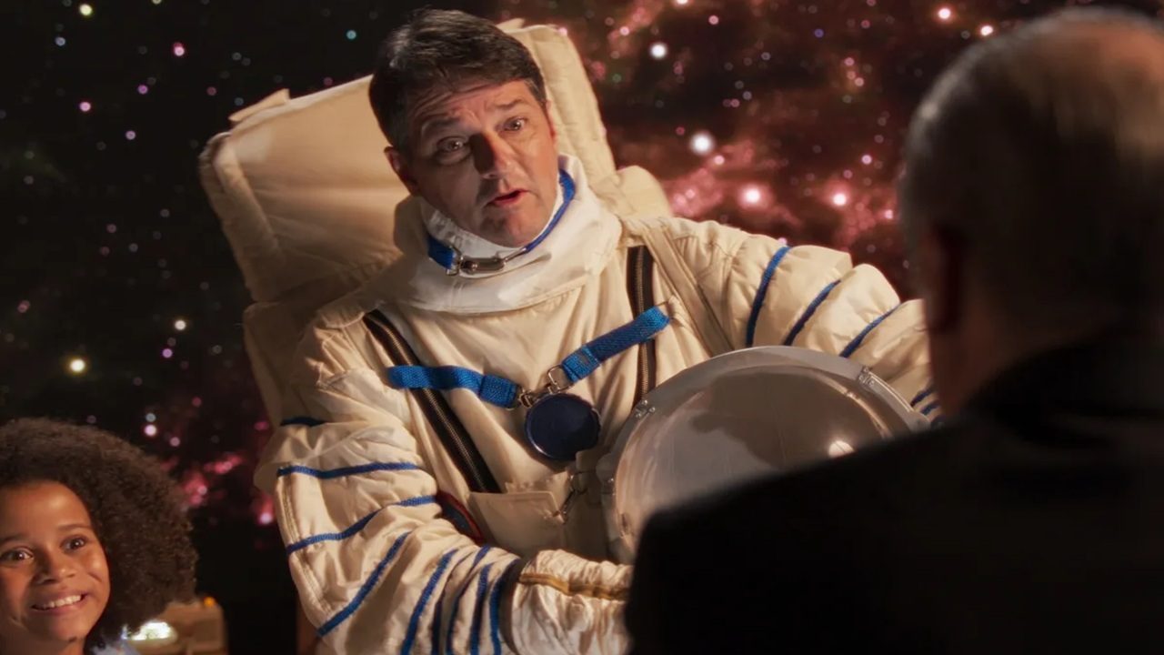 Homem vestido com traje de astronauta segura o capacete enquanto conversa com outra pessoa em um cenário que simula o espaço, com estrelas e nebulosas ao fundo; uma criança observa sorridente ao lado