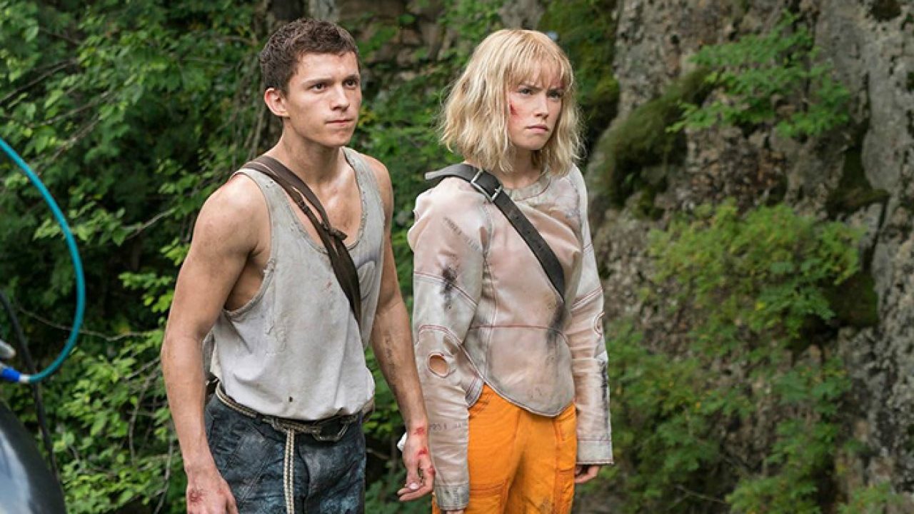 A imagem mostra uma cena do filme "Mundo em Caos" (Chaos Walking), estrelado por Tom Holland e Daisy Ridley. Os personagens aparecem em meio a uma floresta densa, com expressões sérias e atentos ao que está ao redor. Tom veste uma regata branca suja e calça jeans, enquanto Daisy usa uma roupa futurista com tons claros e detalhes em laranja. A atmosfera da imagem sugere tensão e aventura, refletindo o clima distópico da produção.