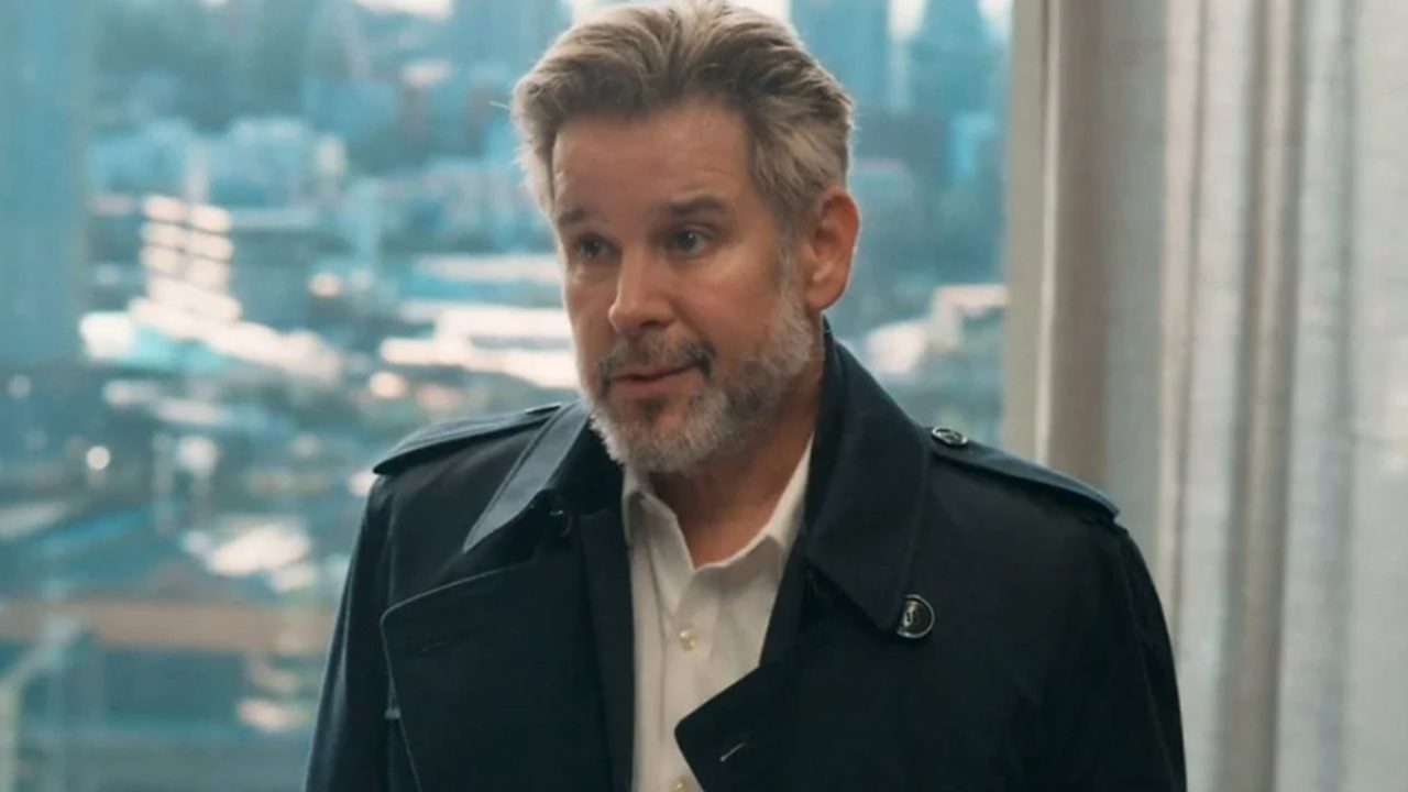 Homem de barba grisalha e casaco escuro conversa em ambiente corporativo com janela e cidade ao fundo