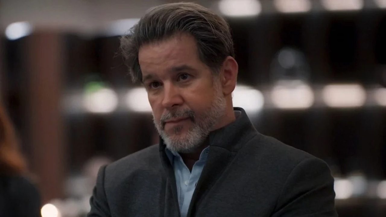 Homem de meia-idade com expressão séria está em um ambiente interno, vestindo camisa azul clara e paletó escuro, com prateleiras desfocadas ao fundo.