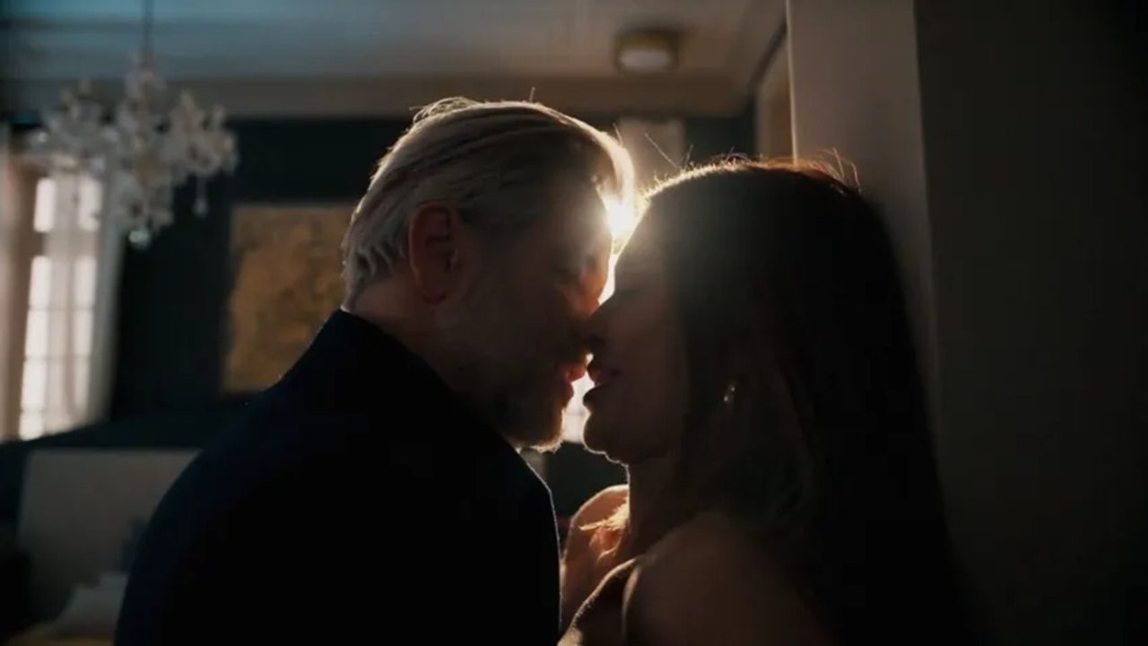 Homem de cabelos grisalhos e mulher se aproximam para beijo em ambiente interno com luz forte ao fundo criando silhuetas em cena dramática