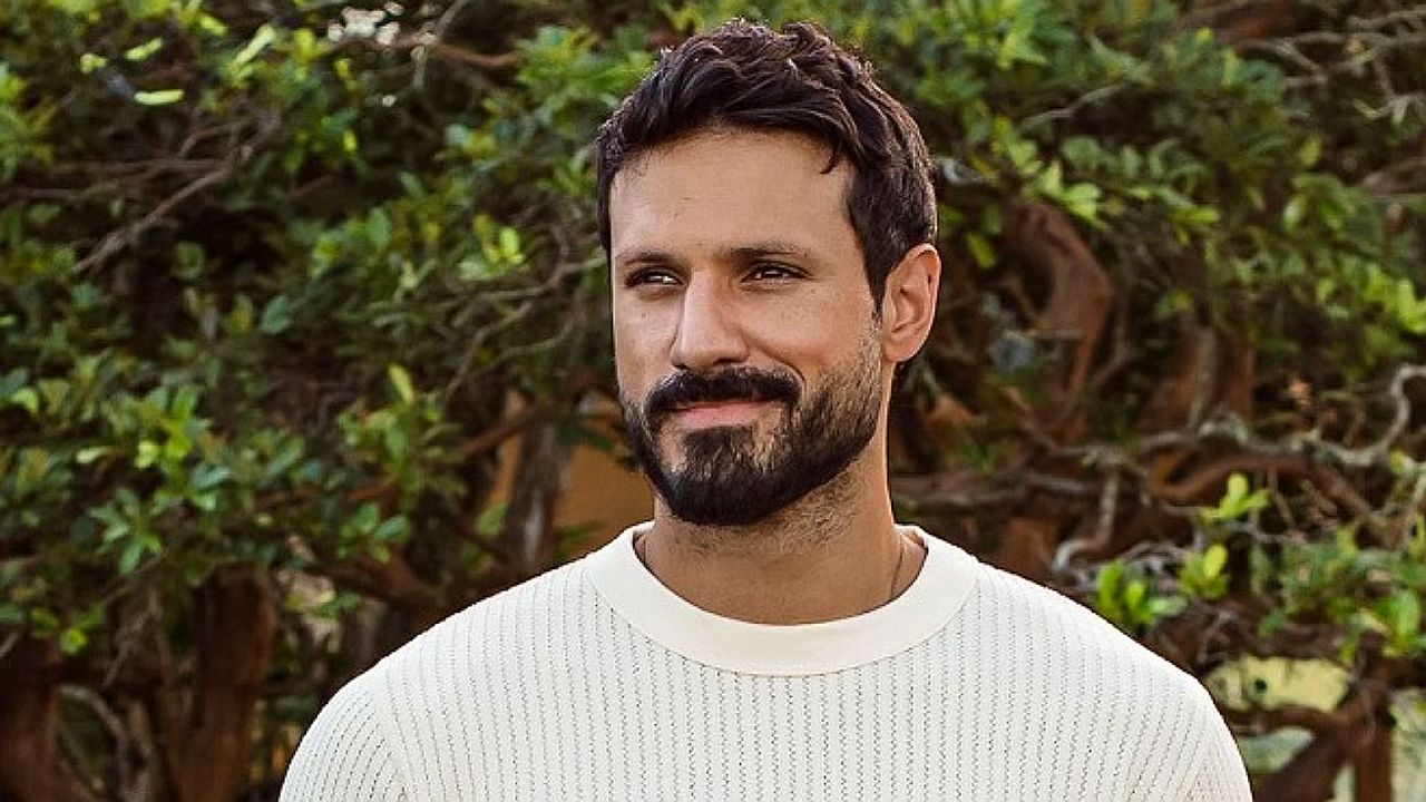 Homem de barba e cabelo escuro sorri levemente enquanto está ao ar livre diante de árvores e vegetação ao fundo
