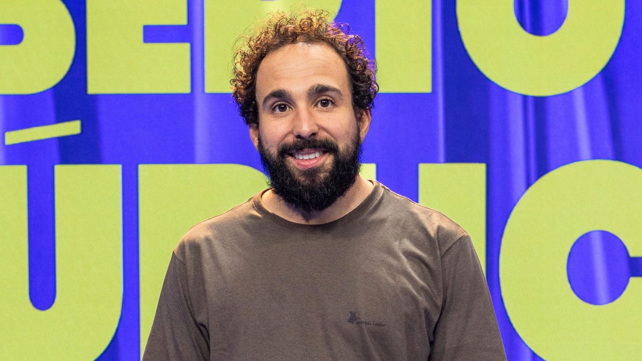 Murilo Couto no palco do programa Aberto ao Público com fundo colorido e letras em destaque