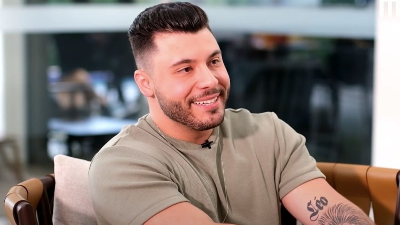 Homem jovem sentado em ambiente interno iluminado, sorrindo enquanto participa de uma conversa. Ele usa camiseta clara, traz barba bem aparada e exibe uma tatuagem no braço direito com o nome “Léo”