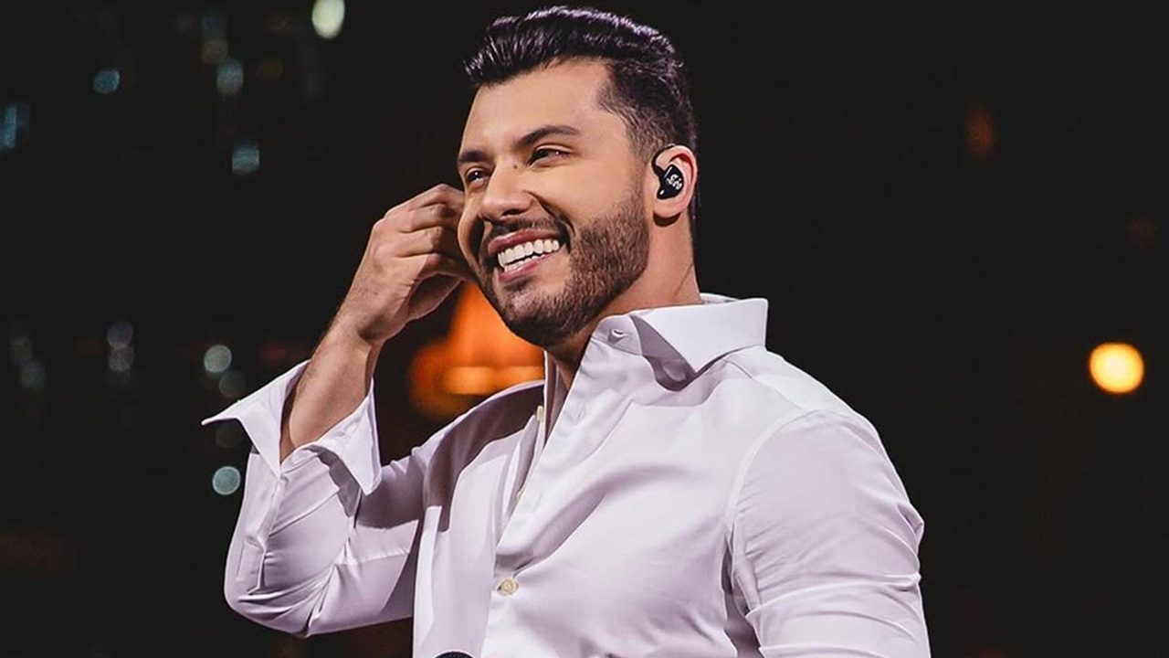 A imagem mostra um homem sorridente usando uma camisa social branca, aparentemente em um palco. Ele segura um ponto eletrônico no ouvido, o que sugere que é um cantor ou apresentador durante uma apresentação. O fundo está escuro, com luzes desfocadas ao longe