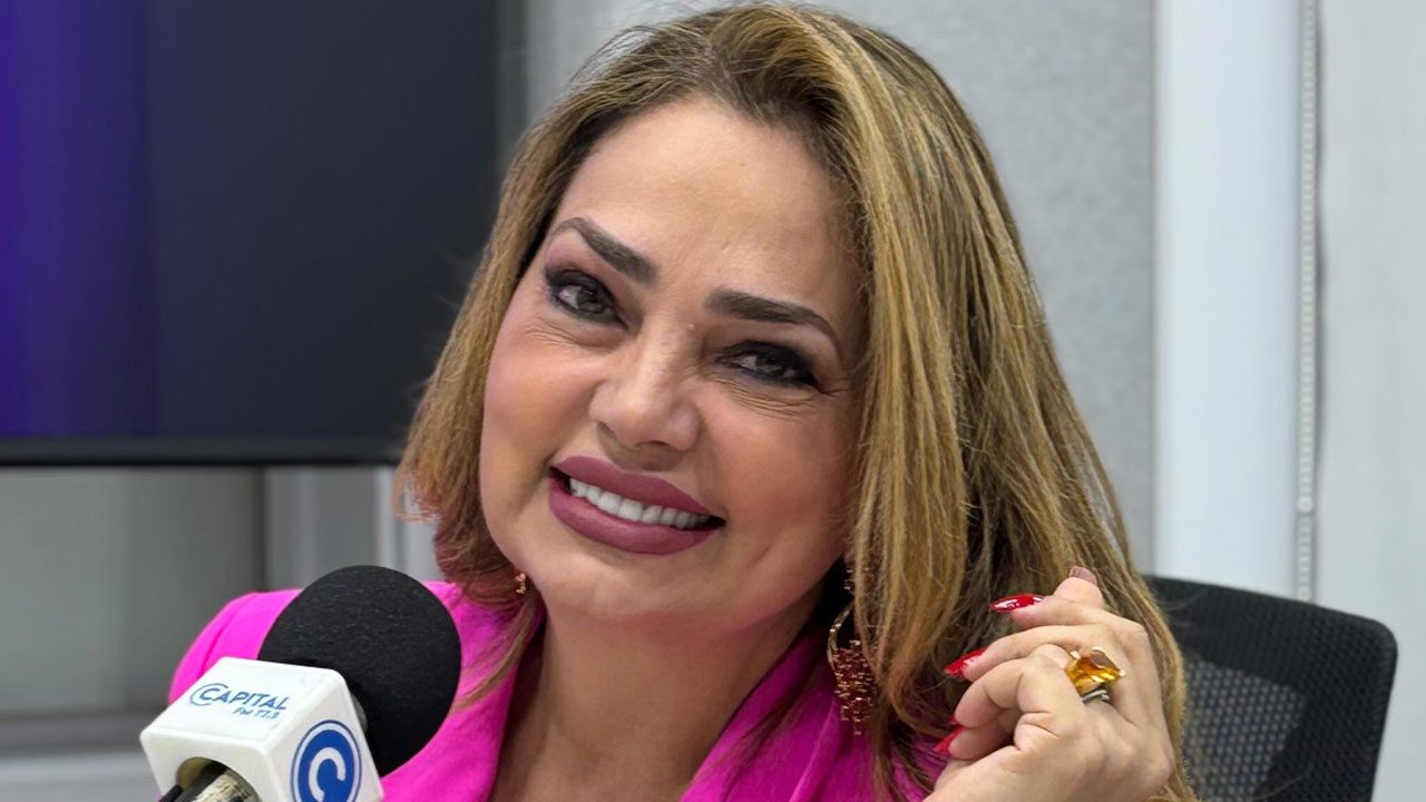 Apresentadora Nani Venâncio sorrindo com blazer pink, maquiagem carregada e cabelo loiro em estúdio da rádio Capital FM