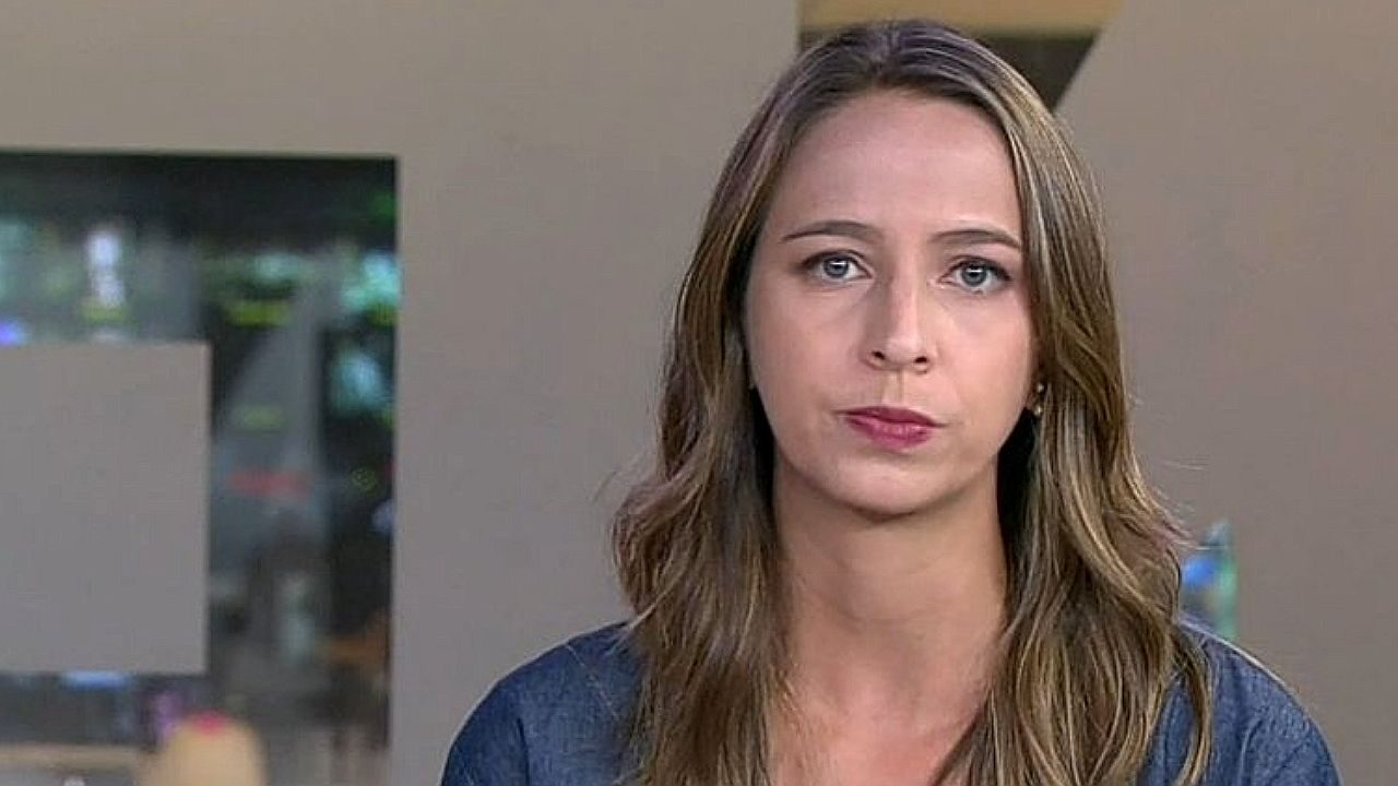 Uma mulher jovem de cabelo castanho claro, comprido e levemente ondulado está olhando diretamente para a câmera com expressão séria. Ela usa uma blusa em tom escuro. A imagem parece ser de uma cena interna, possivelmente em um ambiente doméstico ou escritório, com fundo desfocado que mostra paredes claras e uma abertura ou janela.