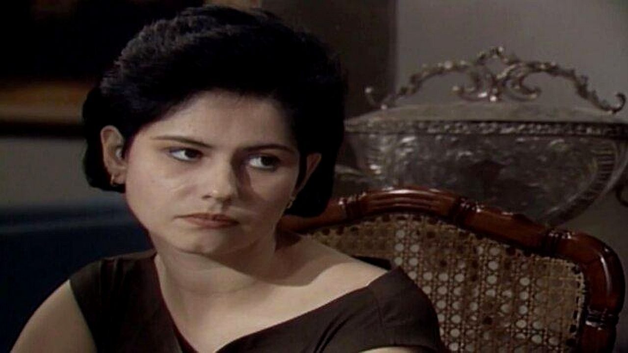 Narjara Turetta com expressão séria sentada à mesa em cena de novela ambientada em sala de estar