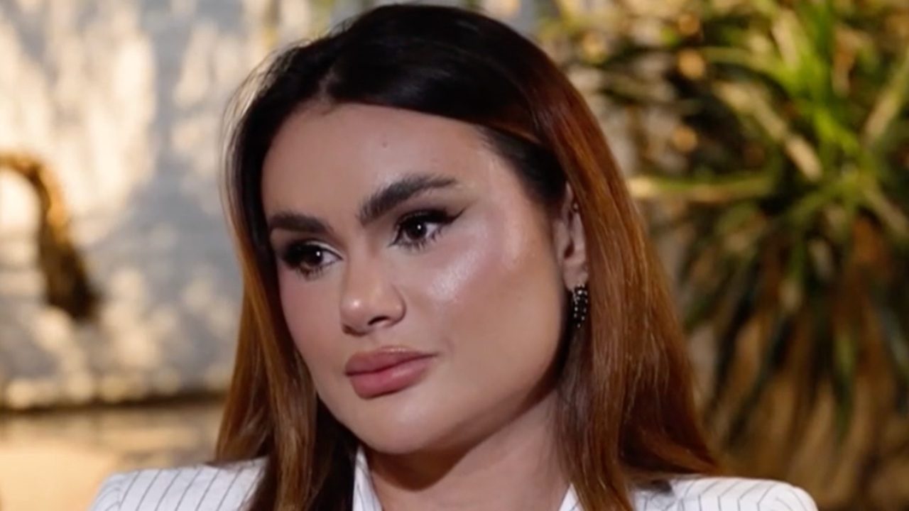 Close-up de uma mulher com expressão séria e olhar pensativo, vestindo blazer claro e maquiagem marcada. O fundo desfocado com plantas sugere um ambiente interno bem iluminado, transmitindo um momento de reflexão ou emoção contida