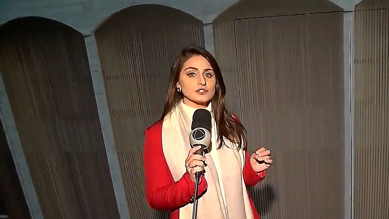 Natália Satler segura microfone da Record TV diante de grandes cilindros metálicos durante gravação de matéria em ambiente industrial