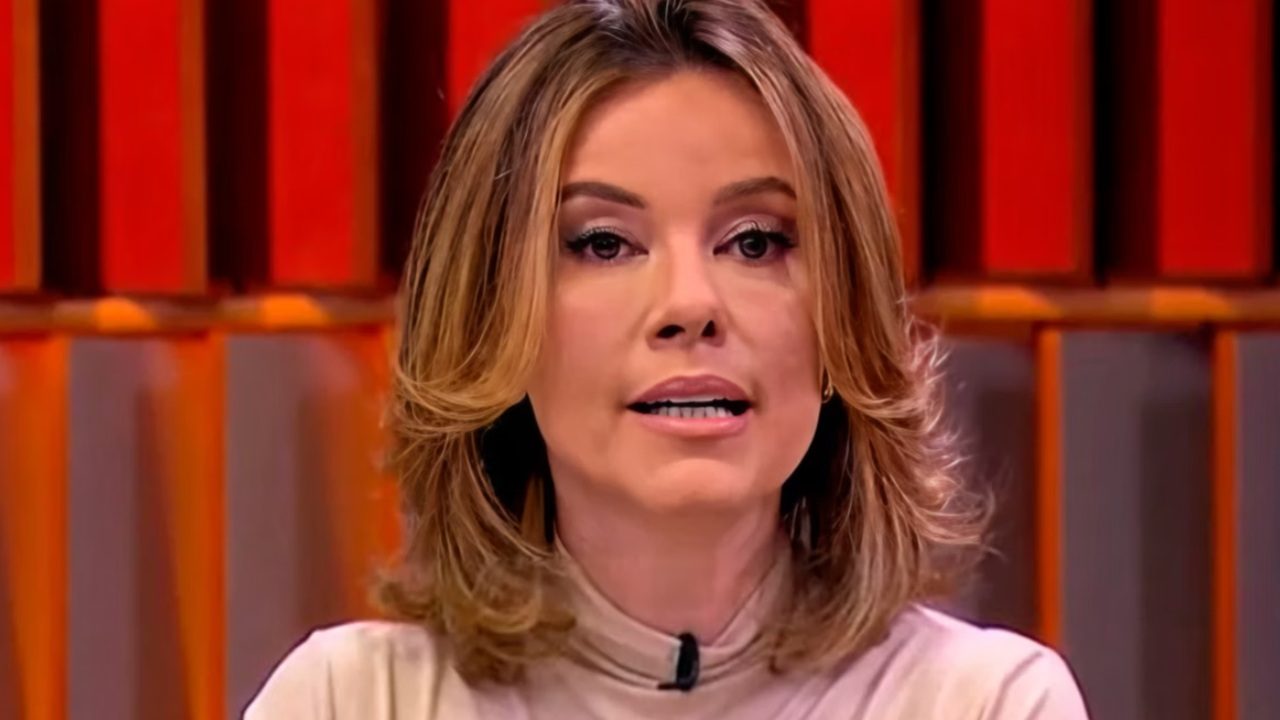 Close de uma mulher apresentando um programa de TV, com expressão séria enquanto fala. Ela usa blusa clara de gola alta e microfone de lapela. Ao fundo, vê-se um cenário iluminado em tons de vermelho e laranja, com linhas verticais desfocadas