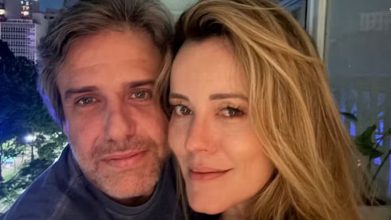 Casal posa junto para selfie em ambiente interno com cidade iluminada ao fundo durante a noite