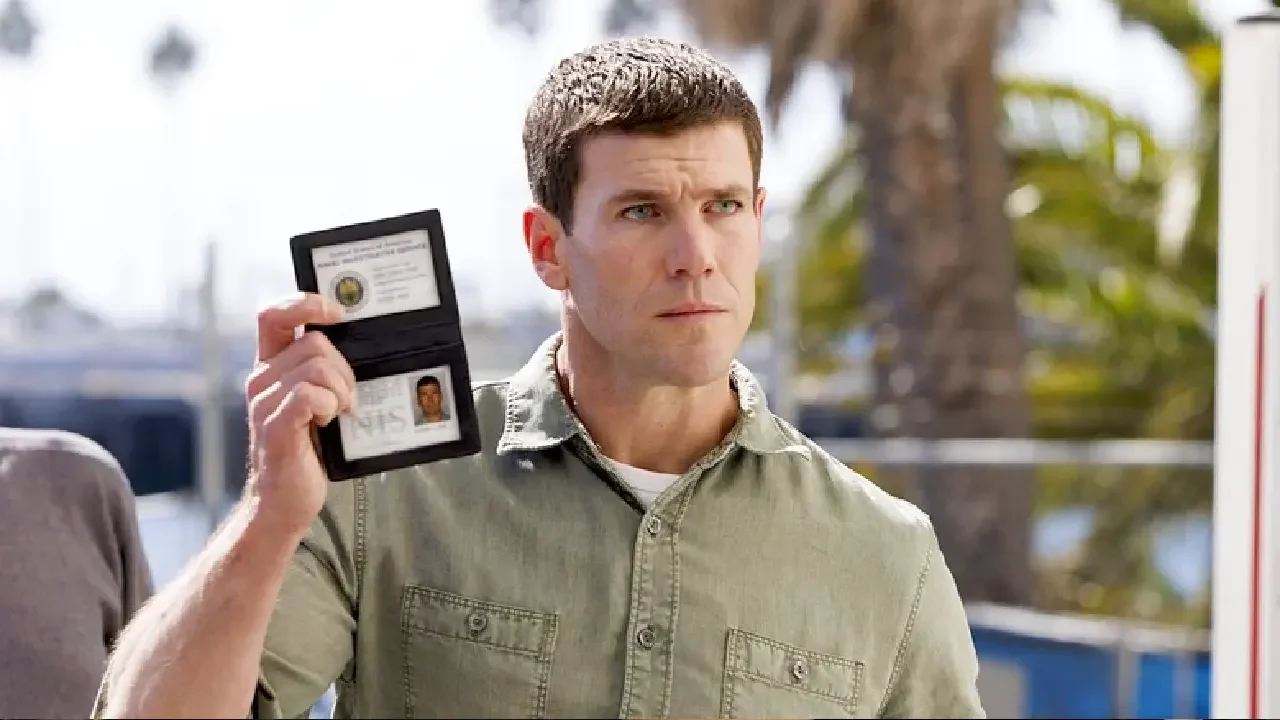 Homem com expressão séria segura uma identificação aberta, mostrando um distintivo e um crachá, sugerindo ser um agente policial ou investigador. Ele usa camisa verde clara e está em um ambiente externo, com palmeiras e luz natural ao fundo.